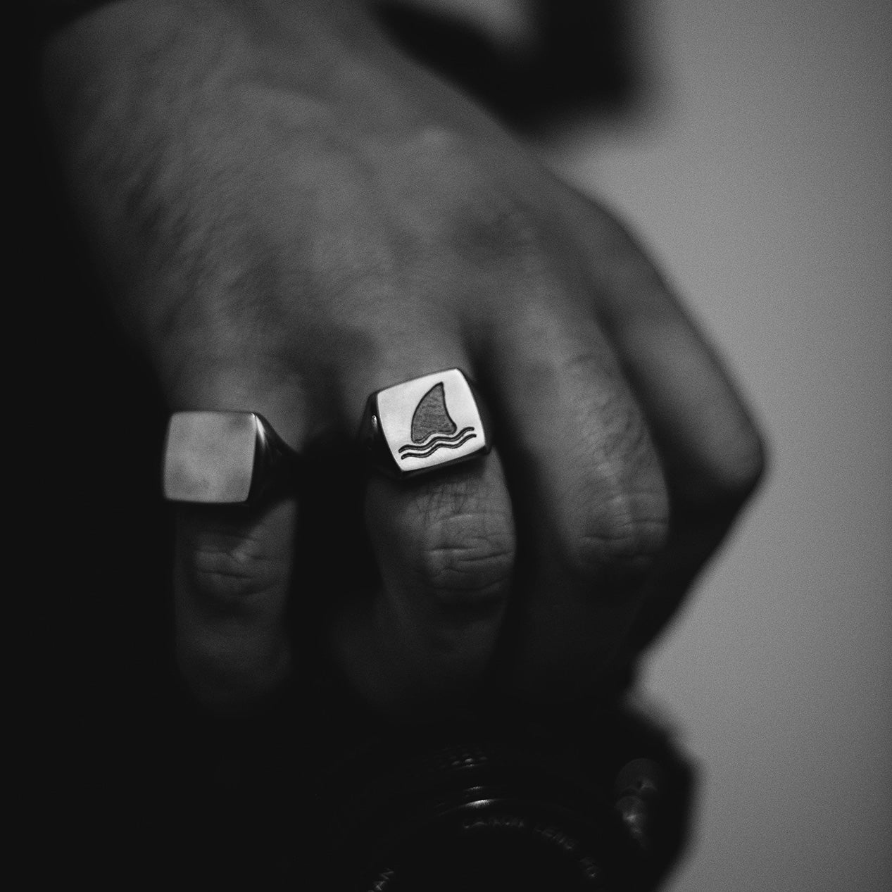 Black and white image of a hand wearing two titanium signet rings, one with no engraving and one with an engraved shark fin.