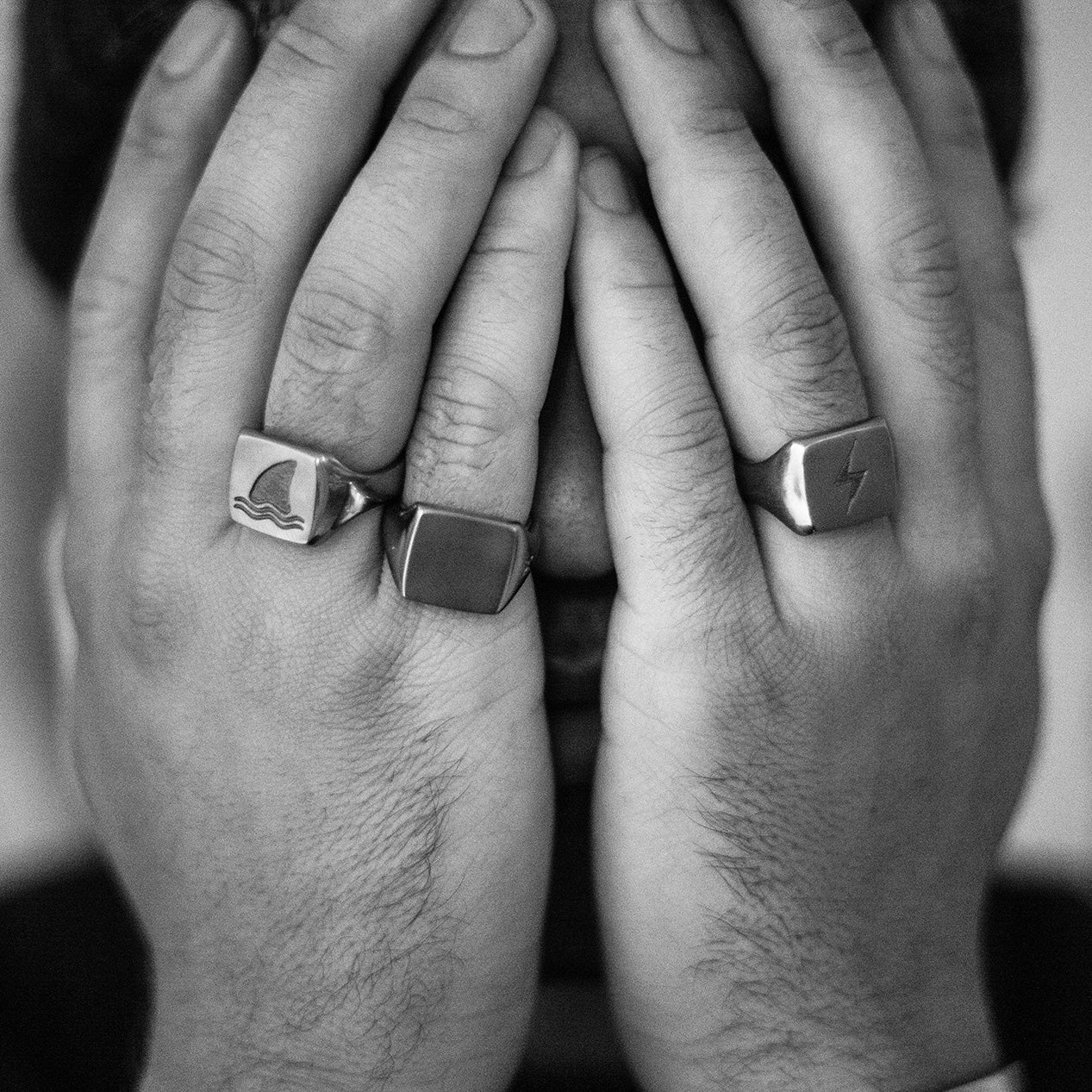 Two hands held in front a face while wearing two titanium signet rings on the left hand and one on the right hand.
