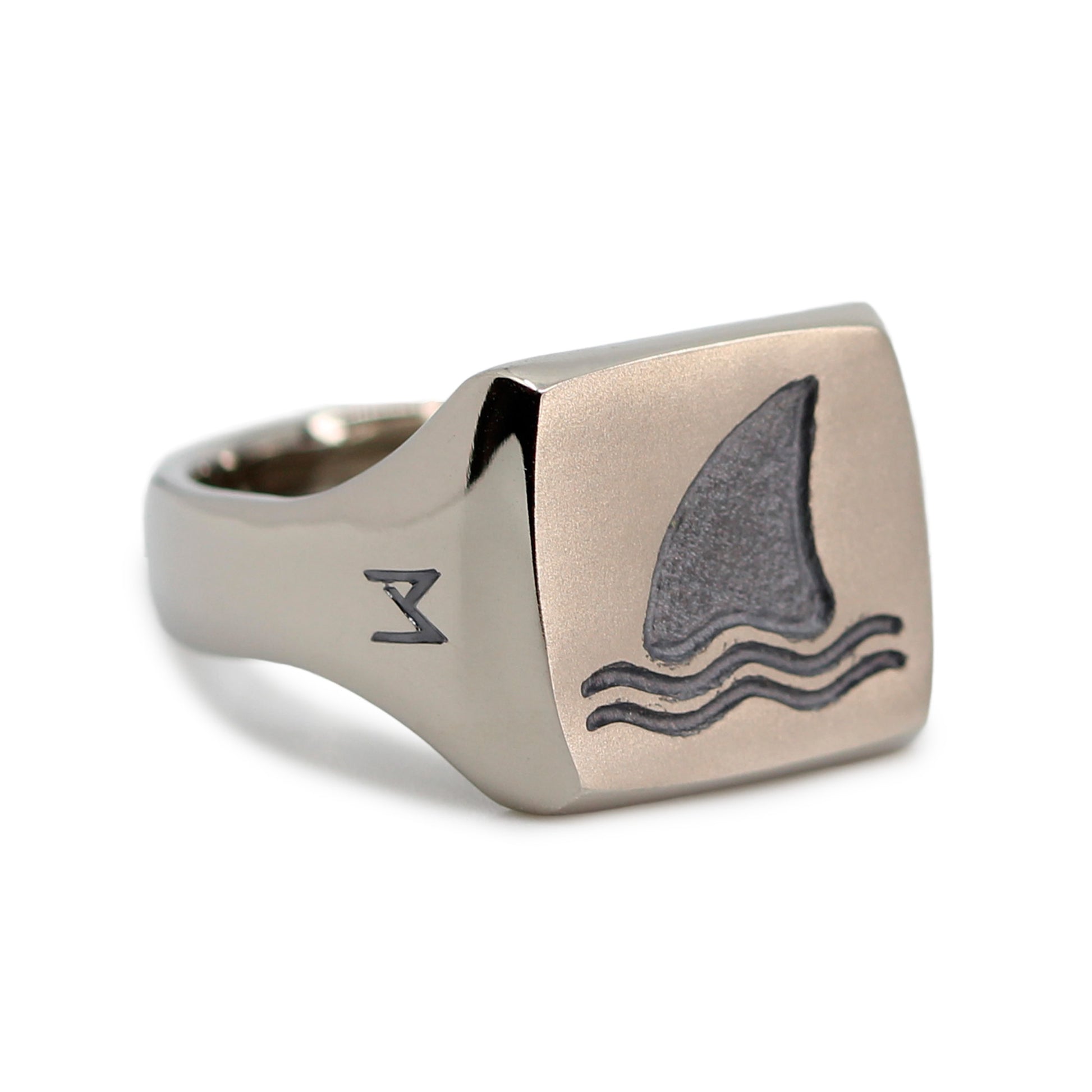 Single silver-colored titanium signet ring, 15x15 mm wide surface with an engraved shark fin. Image shows corner view of the ring