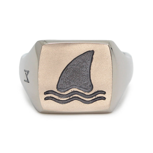 Single silver-colored titanium signet ring, 15x15 mm wide surface with an engraved shark fin. Image shows front view of the ring