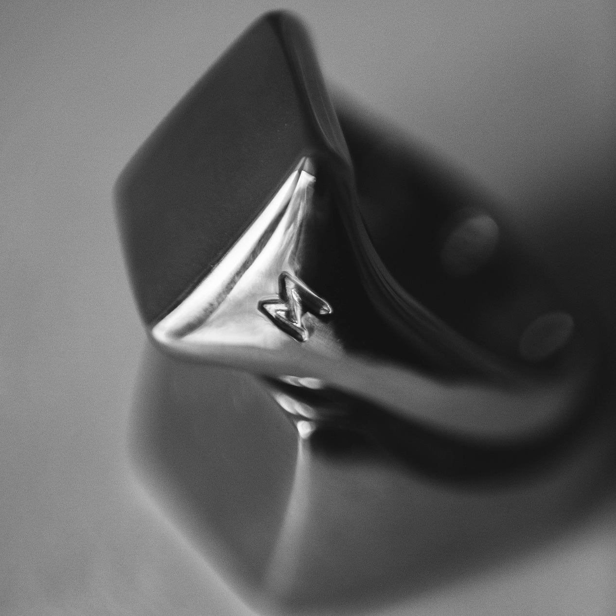 Single silver-colored titanium signet ring, 15x15 mm wide surface. Image shows the ring laying on a reflective surface.