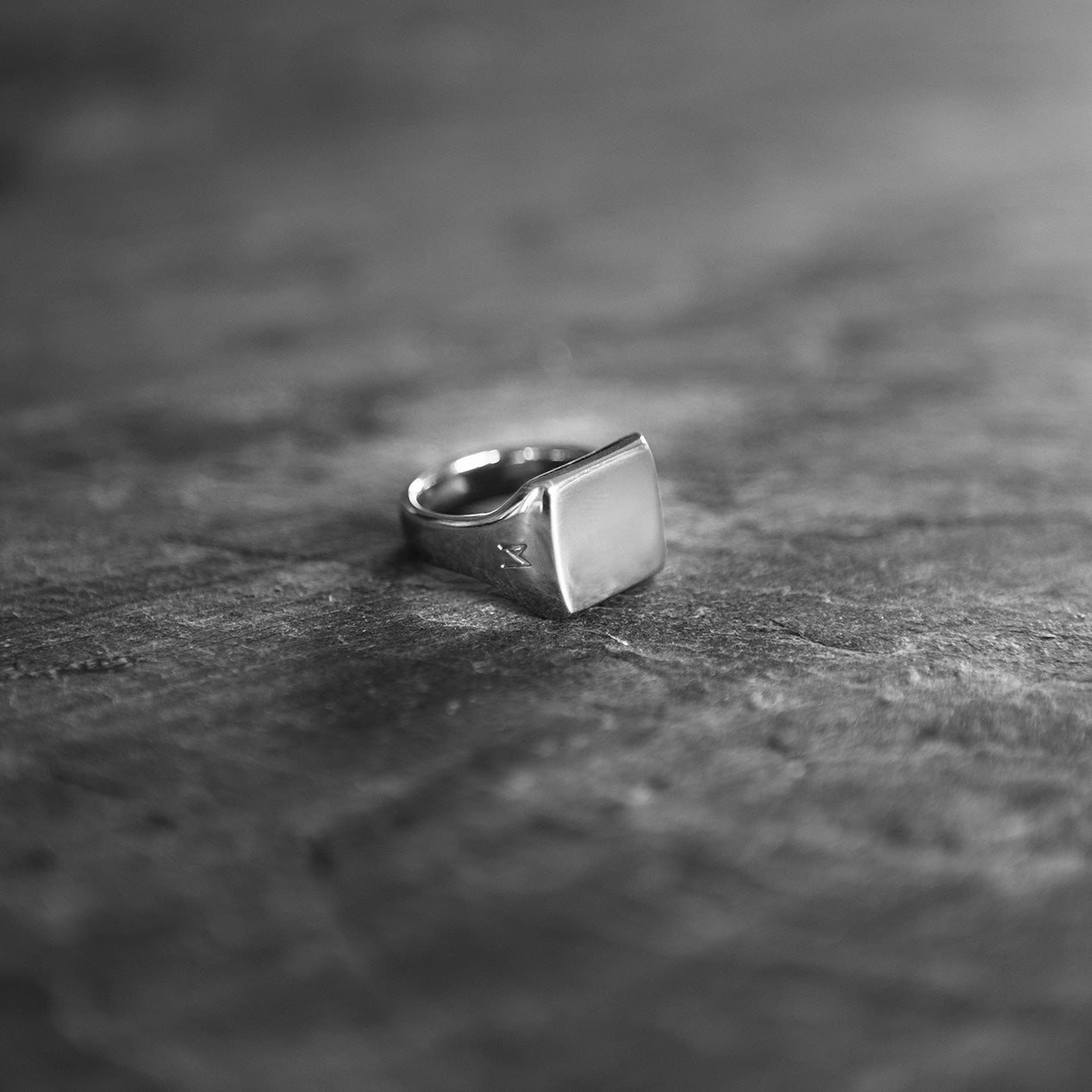 Single silver-colored titanium signet ring, 15x15 mm wide surface. the ring is laying on a rough surface.